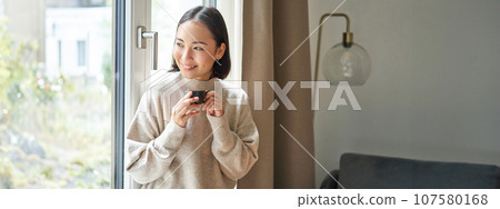 Portrait of beautiful asian woman sitting at home with cup of coffee, enjoying her espresso while looking outside window at passerby, smiling happily Portrait of beautiful asian woman sitting at home with cup of coffee, enjoying her espresso while looking outside window at passerby, smiling happily 107580168