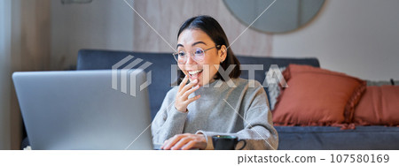 Portrait of asian woman in glasses, sitting with laptop and looking surprised, amazed by promotion on computer 107580169
