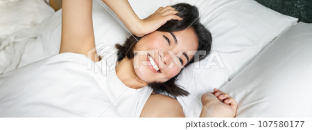 Close up of happy asian girl lying in bed on her pillow, smiling and rolling over as feeling energetic in morning, waking up early 107580177