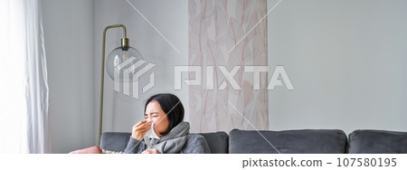 Woman with temperature, coughing, showing symptoms of flu, influenza or cold, sitting in warm clothes on sofa, covered with blanket, sneezing, staying at home in living room 107580195