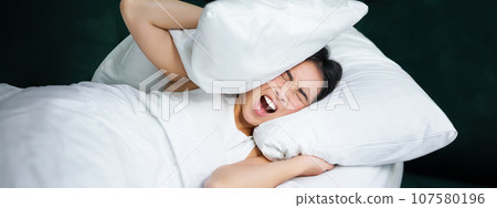 Sad girl with insomnia. Young asian woman lying in bed, cover her ears with pillow, cant sleep, partner snors, loud noise at night disturbing her Sad girl with insomnia. Young asian woman lying in bed, cover her ears with pillow, cant sleep, partner snors, loud noise at night disturbing her 107580196