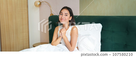 Portrait of excited, smiling asian woman sitting in her bed in morning, daydreaming, looking hopeful 107580210
