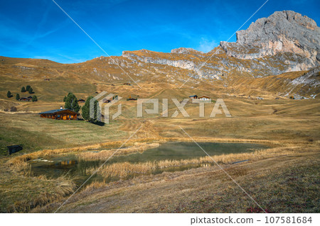 Small lake and wooden lodges on the slope, Dolomites, Italy 107581684