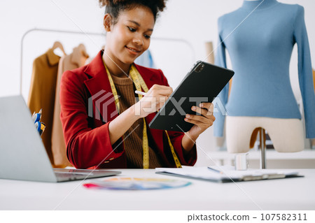 Calm curly brunette dark skinned woman on desk in office of fashion designer and holds tablet and smartphone.. Calm curly brunette dark skinned woman on desk in office of fashion designer and holds tablet and smartphone.. 107582311