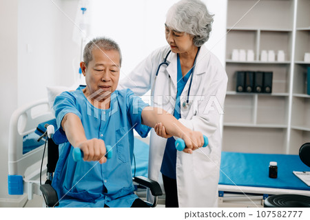 Asian physiotherapist helping elderly man patient stretching arm during exercise correct with dumbbell in hand during training hand in bed in clinic Asian physiotherapist helping elderly man patient stretching arm during exercise correct with dumbbell in hand during training hand in bed in clinic 107582777