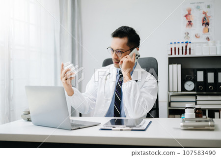 Asian Doctor talking over his mobile phone and making notes in a note. Medicine professional talking on phone and clinical report test results. 107583490