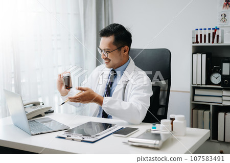 Confident Young Asian male doctor in white medical uniform sit at desk working on computer. Smiling use laptop write in medical journal in clinic. Confident Young Asian male doctor in white medical uniform sit at desk working on computer. Smiling use laptop write in medical journal in clinic. 107583491
