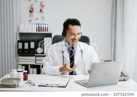 Attractive male doctor talking while explaining medical treatment to patient through a video call with laptop in office 107583504