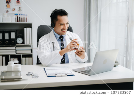Attractive male doctor talking while explaining medical treatment to patient through a video call with laptop in office 107583507