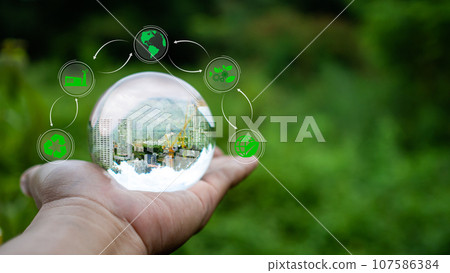 Concept of renewable energy, environmental protection, and sustainable renewable energy sources. An image of a building under construction in crystal glass resting on a person's hand and with a 107586384