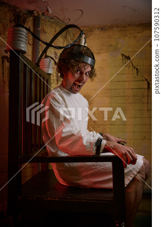 Crazy man screaming feeling madness on electric chair at psychiatric hospital 107590312