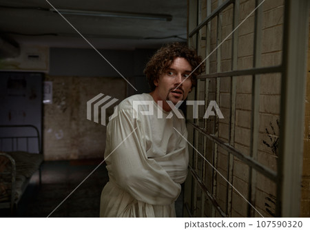 Scared quiet crazy man wearing straightening jacket standing nearby cell gate 107590320