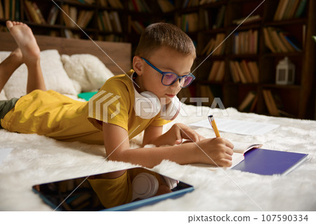 Little boy child using modern gadget studying at home lying in bed Little boy child using modern gadget studying at home lying in bed 107590334