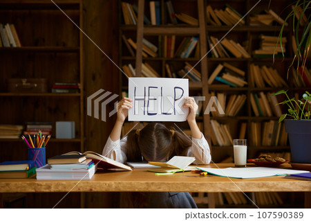 Tired girl student lying on home desk holding placard with help word Tired girl student lying on home desk holding placard with help word 107590389