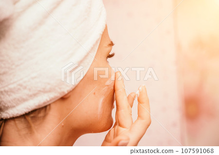 Young beautiful woman using face cream moisturizing lotion after bath. Pretty attractive girl in a towel on her head stands in front of a mirror in a home bathroom. Daily hygiene and skin care 107591068