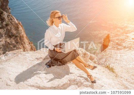 Business woman on nature in white shirt and black skirt. She works with an iPad in the open air with a beautiful view of the sea. The concept of remote work. Business woman on nature in white shirt and black skirt. She works with an iPad in the open air with a beautiful view of the sea. The concept of remote work. 107591099