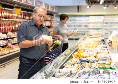 Mature man diligently choosing fresh cheese in supermarket Mature man diligently choosing fresh cheese in supermarket 107591607