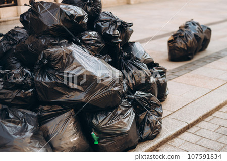 Pile of black garbage on the footpath at side road in big city, pollution trash. Many garbage plastic bags black waste, pollution, lots junk dump 107591834