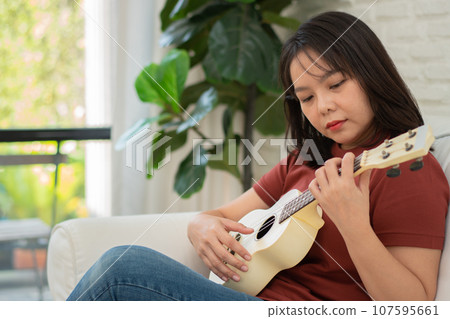 Happy Asian woman sitting on sofa in living room and practicing ukulele in leisure. Relaxation and inspiration from artist. Concept of music, relax and hobby activity enjoy in leisure Happy Asian woman sitting on sofa in living room and practicing ukulele in leisure. Relaxation and inspiration from artist. Concept of music, relax and hobby activity enjoy in leisure 107595661