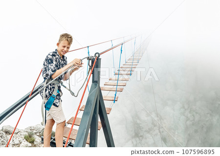 Boy fastens safety belt carabine to walk along rope bridge 107596895