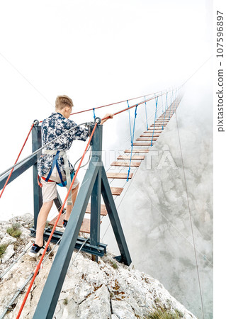 Brave boy walks along rope bridge in foggy mountain park Brave boy walks along rope bridge in foggy mountain park 107596897