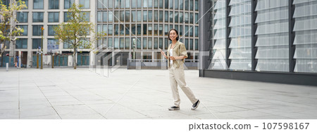 Portrait of beautiful smiling asian woman, walking on street of city centre with smartphone, looking at camera Portrait of beautiful smiling asian woman, walking on street of city centre with smartphone, looking at camera 107598167