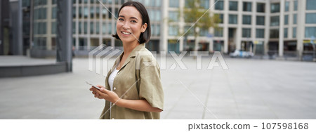Portrait of asian woman standing on street, city square and holding mobile phone. Girl with smartphone walking outdoors Portrait of asian woman standing on street, city square and holding mobile phone. Girl with smartphone walking outdoors 107598168