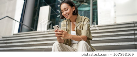 Technology and communication. Young smiling girl, asian woman sits with smartphone, reads message with big smile Technology and communication. Young smiling girl, asian woman sits with smartphone, reads message with big smile 107598169