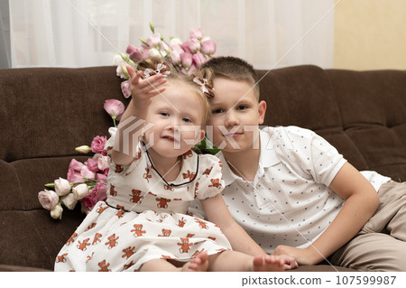 Family concept. Brother and sister play happily while sitting on a brown sofa, in a home interior against the background of a pink bouquet of flowers. Love and interaction. 107599987