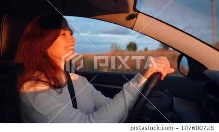 Cheerful young woman drives car along rural road at sunset light. Smiling woman drives car holding steering wheel across countryside in evening. Woman driver enjoys car trip across farmland 107600728