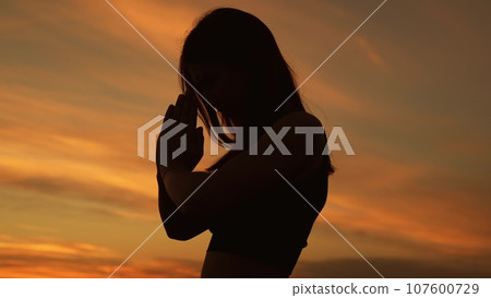Black silhouette of praying woman against sunset sky with light clouds. Woman puts hands in pray gesture sending wishes to evening sky. Religious woman prays outdoors at sunset light Black silhouette of praying woman against sunset sky with light clouds. Woman puts hands in pray gesture sending wishes to evening sky. Religious woman prays outdoors at sunset light 107600729