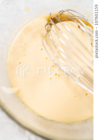Mixing wet and dry ingredients with a hand whisk in a glass mixing bowl to bake a carrot bundt cake with cream cheese frosting. 107601159