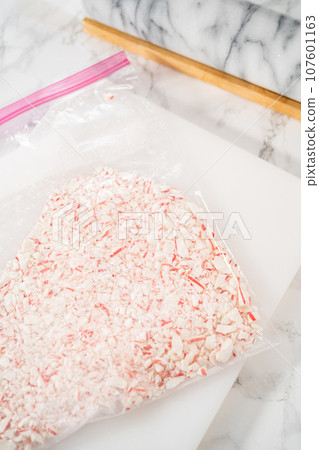 Crushing peppermint candies in a plastic bag with a marble rolling pin on a white cutting board. 107601163