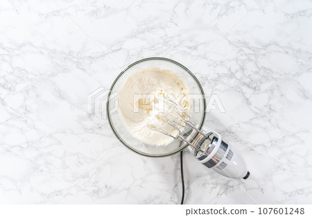 Flat lay. Whisking buttercream frosting in a glass mixing bowl with an electric hand mixer to prepare the peppermint buttercream frosting. 107601248