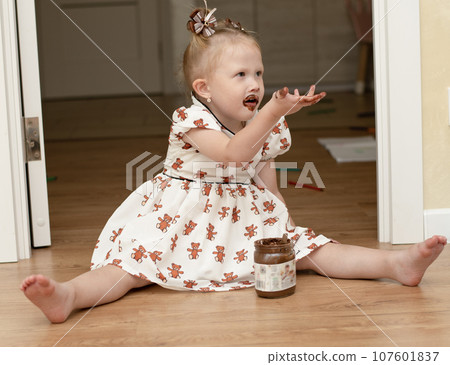 A little girl 3 years old in a beautiful white dress sits on the floor in a home interior and happily eats chocolate spread from a jar. His whole mouth is ridiculously smeared with chocolate. 107601837