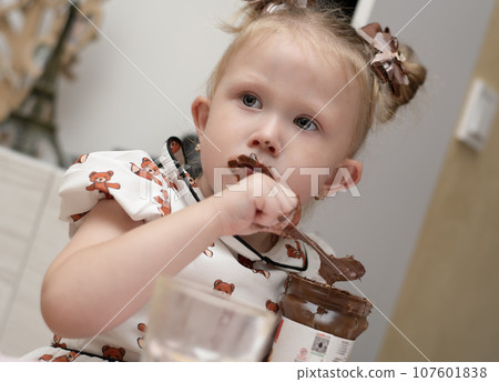 A little girl 3 years old in a beautiful white dress sits on the floor in a home interior and happily eats chocolate spread from a jar. His whole mouth is ridiculously smeared with chocolate. 107601838