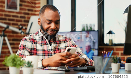 Happy cheerful person chatting with online friends over the phone, enjoying himself in welcoming stylish home. Entertained african american man scrolling on his social media feed, handheld camerawork 107604223
