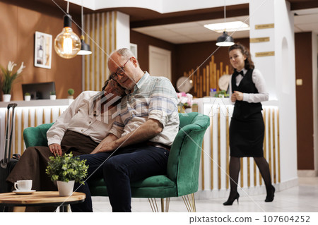 Exhausted elderly couple snooze on nice couches in luxurious hotel lobby. After long journey, retired senior vacationers resting in lounge area while waiting for check-in. Friendly resort employees. 107604252