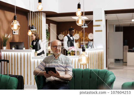 Caucasian senior person sits on hotel lobby sofa, holding digital tablet. Old man waiting in luxurious lounge area and uses smart device for browsing on internet. Luxurious and relaxing atmosphere. 107604253