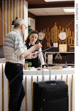 Senior man tourist checks in at the front desk of a hotel, eager for a pleasant holiday. While filling out booking reservation documents, a kind female concierge advises an old man traveller. 107604254