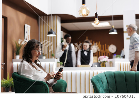 Caucasian senior woman enjoying vacation, browsing on smartphone. Friendly receptionists assists client with check-in at modern hotel lobby. Emphasis on hospitality and technology. 107604315