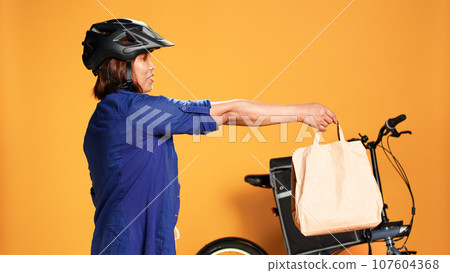 BIPOC food delivery worker reaching order address, waiting for customer to answer door. Courier woman greeting client, offering takeaway lunch bag, isolated over orange studio background BIPOC food delivery worker reaching order address, waiting for customer to answer door. Courier woman greeting client, offering takeaway lunch bag, isolated over orange studio background 107604368