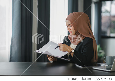 Young muslim millennial businesswoman in brown hijab headscarf working with financial reports while sitting in modern office. Young muslim millennial businesswoman in brown hijab headscarf working with financial reports while sitting in modern office. 107605955