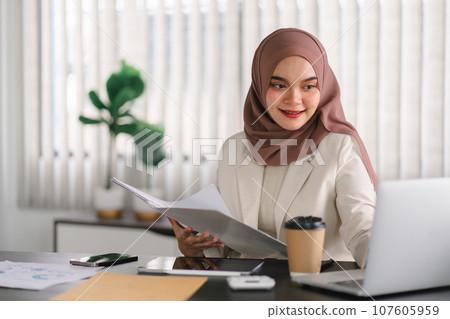 Young muslim millennial businesswoman in brown hijab headscarf working with financial reports while sitting in modern office. Young muslim millennial businesswoman in brown hijab headscarf working with financial reports while sitting in modern office. 107605959