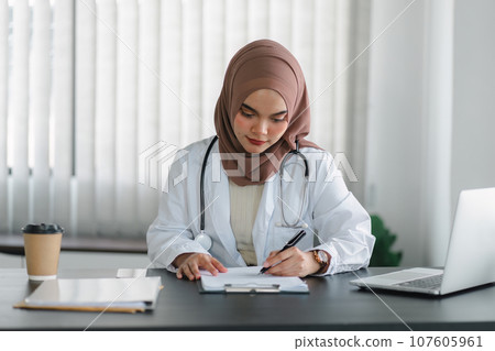 Young muslim female doctor in brown hijab headscarf working with medical papers while sitting in modern clinic office. 107605961