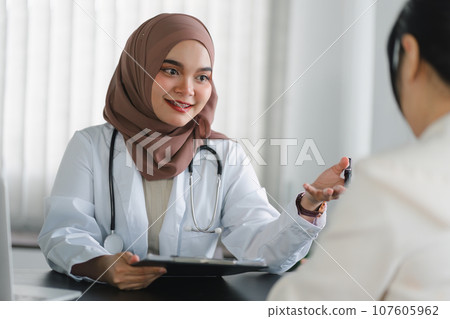 Young muslim female doctor in brown hijab headscarf having appointment consulting female patient in modern clinic hospital. Medical healthcare concept. 107605962