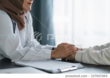 Young muslim female doctor in brown hijab headscarf holding hands with woman for hope and kindness at modern clinic office. 107605969