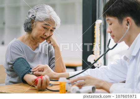 A young doctor using digital tonometer check blood pressure for senior woman. A doctor is holding a senior woman's hand. A young doctor using digital tonometer check blood pressure for senior woman. A doctor is holding a senior woman's hand. 107605971
