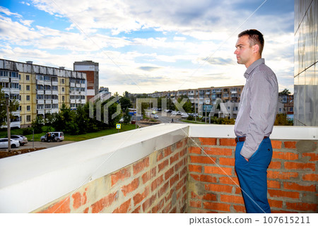Man in office clothes standing on the balcony and looking at the old dirty Russian city of Bolshoy Kamen. He is sad and serious. depressive concept of loneliness, failure, loss of money Man in office clothes standing on the balcony and looking at the old dirty Russian city of Bolshoy Kamen. He is sad and serious. depressive concept of loneliness, failure, loss of money 107615211