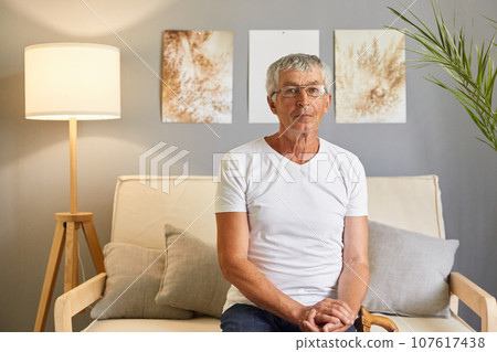 Serious confident grey-haired mature man wearing casual white T-shirt and glasses sitting on sofa at home interior resting at home on couch looking at camera with concentrated expression. Serious confident grey-haired mature man wearing casual white T-shirt and glasses sitting on sofa at home interior resting at home on couch looking at camera with concentrated expression. 107617438